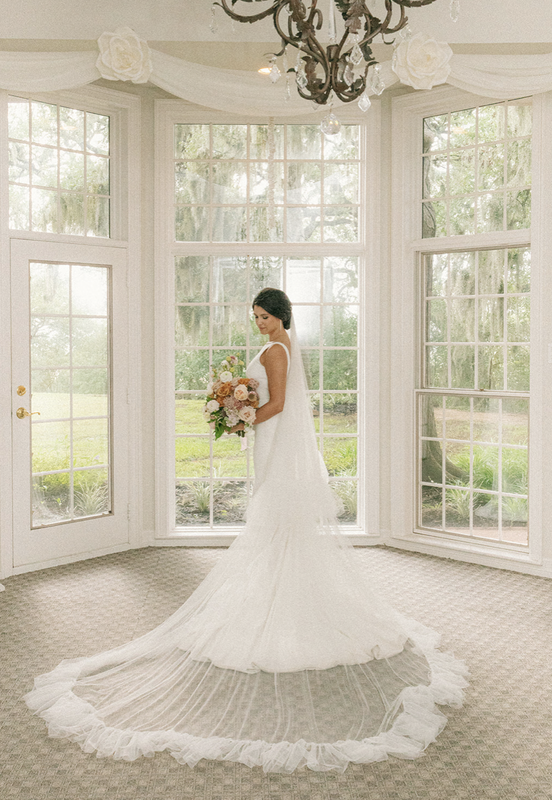 Bride with long train and pink flowers with beautiful windows in the background overlooking hill country.